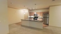 This single-floor space showcases an open-concept layout that integrates the kitchen and living area into one cohesive environment. The kitchen serves as the focal point, anchored by a large center island topped with a dark countertop and fitted with a stainless steel double sink. Overhead, pendant lights hang at varying heights, providing focused task lighting above the island and enhancing the kitchen's visual appeal. A track lighting system further illuminates the space.

Cabinetry in a warm medium-toned wood finish lines the kitchen walls, offering generous storage. Stainless steel appliances, including a refrigerator and microwave, are complemented by a black range, creating a contemporary contrast. The countertops throughout carry a consistent dark tone, adding definition to the lighter cabinetry.

The adjoining living area features neutral wall-to-wall carpeting, providing a soft transition from the hard flooring of the kitchen. The warm, neutral color palette maintained throughout creates a cohesive and inviting ambiance. The open floor plan allows for flexible furniture arrangement and promotes a sense of spaciousness throughout the unit.