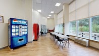 This single-floor space functions as a multipurpose community or common room, designed to accommodate a variety of activities and social gatherings. The interior features light hardwood-style flooring throughout, neutral cream-colored walls, and a drop ceiling with recessed panel lighting that ensures consistent, even illumination across the room.

The main area is generously proportioned, with notably tall ceilings that contribute to an open, airy atmosphere. Long folding tables with stackable chairs are arranged to support group meetings or communal dining, while a separate seating arrangement with a round table offers a more casual setting. A row of red upholstered chairs near the entrance provides additional seating.

Large floor-to-ceiling windows fitted with roller shades line one wall, allowing ample natural light and offering views of a landscaped outdoor area with benches and greenery. A glass exit door provides direct outdoor access. Pepsi and Coca-Cola beverage vending machines, along with a snack vending machine, are conveniently positioned within the space. A wooden bookshelf stocked with reading materials adds a practical touch. Decorative elements include potted plants and wall artwork, enhancing the overall welcoming ambiance.