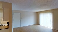 This single-floor space presents a clean and functional layout centered around an open living area. The room features light wood-style laminate flooring that extends throughout, lending warmth and a sense of continuity to the space. Neutral beige walls provide a blank canvas adaptable to a variety of interior styles.

A textured popcorn ceiling adds dimension overhead, while a solid white entry door with a standard deadbolt serves as the main point of entry. On the opposite side of the room, a large window dressed with vertical blinds allows for generous natural light to filter in, brightening the interior and creating an airy atmosphere.

The overall layout is straightforward and open, offering flexible furniture placement options. The space maintains a modest and practical character, with minimal architectural embellishment, making it well-suited for those seeking a move-in ready environment with a neutral aesthetic foundation. The single-floor design ensures ease of accessibility and a seamless flow throughout the living area.