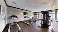Located at 1175 Riverwalk Terr in Jenks, Oklahoma, this single-floor fitness and recreation facility offers a well-appointed space designed to support an active lifestyle. The exercise room is generously sized and outfitted with a comprehensive selection of cardio and strength equipment, including multiple treadmills, elliptical machines, a multi-station cable machine, free weights, and weight benches — all from recognized brands such as Precor. Rubber flooring throughout the exercise areas provides durability and safety, while recessed lighting and a wall-mounted flat-screen TV enhance the workout environment. Large, multi-pane windows trimmed in dark wood frame outdoor views, filling the space with natural light. A dedicated recreation area features soaring coffered ceilings with rich dark crown molding, arched transom windows, and French doors that open to an outdoor patio. A ping pong table serves as a central gathering point in this room, which also includes a built-in kitchenette with dark cabinetry and granite countertops. Four connecting hallways provide seamless circulation throughout the facility. The overall design blends classic architectural detailing with functional, modern amenities, creating an inviting and well-equipped community fitness destination.