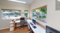 This compact, single-floor business center is thoughtfully designed to support a productive work environment. The space is anchored by a row of sleek, white built-in desks running along two walls, each outfitted with computer workstations and ergonomic rolling chairs. Large, wood-trimmed windows line the walls and offer generous views of lush, landscaped greenery outside, flooding the interior with natural light and creating a calm, focused atmosphere.

The flooring throughout is warm hardwood, lending a clean and professional feel to the space. Overhead fluorescent strip lighting supplements the natural light, ensuring consistent illumination across all work areas. A dedicated support zone features a white multi-drawer filing cabinet topped with a multifunction printer, alongside a distinctive teal accent wall adorned with framed textile artwork, adding a touch of visual character to the functional layout.

A glass-partitioned enclosure — likely a private office or conference nook — is visible through one wall, accessible via a tiled entryway and providing an option for more private meetings or conversations. Decorative touches such as potted plants and framed art pieces, including a notable Andy Warhol print, contribute to an inviting and creative work setting. The overall design balances practicality with comfort, making it a well-equipped and pleasant shared workspace.