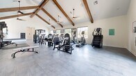 Located at 3800 Centerplace Drive in Greeley, Colorado, this fitness center offers a generously sized, single-floor workout facility designed to support a wide range of exercise routines. The space is immediately distinguished by its soaring vaulted ceilings accented with decorative exposed wood beams, creating an open and energizing atmosphere. Ceiling fans are positioned throughout to maintain comfortable airflow during workouts.

The main workout floor is finished with durable large-format tile flooring in a neutral gray tone, providing a clean and modern aesthetic. Abundant natural light streams in through multiple windows along the walls, complementing the bright white interior and keeping the space feeling open and inviting. Wall-mounted televisions are positioned for easy viewing from cardio equipment.

The cardio section is well-equipped with multiple treadmills, elliptical trainers, stationary bikes, a rowing machine, and a stair climber. The strength training area features a comprehensive free weights section with a full rack of dumbbells, a multi-station cable machine, a power rack with barbells and bumper plates, and various resistance machines targeting different muscle groups. Functional training accessories, including medicine balls, battle ropes, and stability balls, are neatly organized on dedicated racks. Full-length mirrors line portions of the walls, enhancing the sense of space while allowing users to monitor their form.