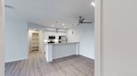 This single-floor space showcases a clean, contemporary design centered around an open-concept layout that seamlessly connects the kitchen and living areas. Light hardwood flooring runs throughout, complementing the soft, neutral wall tones that lend an airy, welcoming ambiance to the interior.

The kitchen is a standout feature, equipped with stainless steel appliances including a refrigerator and microwave. White cabinetry provides ample storage, while a peninsula with a built-in breakfast bar offers additional workspace and casual seating. Pendant lighting hangs above the peninsula, adding a modern, stylish touch, and recessed lighting throughout ensures the space remains well-illuminated.

The open living area is served by a ceiling fan with sleek, modern blades, contributing both function and visual appeal. A walk-in pantry or storage closet is visible toward the back, providing practical organizational space. The overall design emphasizes simplicity and functionality, with a bright, neutral palette and modern fixtures that create a cohesive and inviting atmosphere throughout the unit.