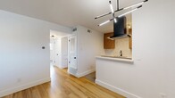 This well-appointed 2-bedroom, 1-bathroom residence offers a clean, modern aesthetic defined by white walls, warm hardwood-style flooring, and contemporary finishes throughout. The open-concept kitchen and living area serves as the heart of the home, where a stylish peninsula with a white quartz countertop and a built-in gas cooktop anchors the space. The kitchen is equipped with rich wood-toned cabinetry, a white tile backsplash, stainless steel appliances including a refrigerator and dishwasher, a matte black single-basin sink, and a striking black geometric pendant light fixture overhead. A sleek wall-mounted range hood adds a bold design accent. Both bedrooms feature the same warm hardwood-style flooring, crisp white walls, and natural light from windows fitted with horizontal blinds. The spacious primary bedroom includes a ceiling fan, while the second bedroom offers a similarly bright and airy feel. The bathroom is finished with a wood-toned vanity, quartz countertop, a large mirror, and a tub-shower combination accented by dramatic marble-style tile surround. Two hallways and a dedicated closet provide practical storage and circulation throughout the unit.
