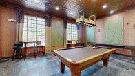 Located in Humble, TX, this dedicated game room is a well-appointed entertainment space designed for leisure and social gatherings. The room is anchored by a solid wood billiard table at its center, complemented by a full-length shuffleboard table along one wall, making it an ideal venue for recreational activities. A wall-mounted flat-screen TV adds a modern entertainment option to the space.

The design aesthetic blends elegance with warmth. The ceiling is a standout architectural feature, adorned with ornate embossed tin tiles framed by rich dark wood trim, lending a classic, upscale character to the room. A statement pendant light fixture with amber-toned shades hangs above the billiard table, providing focused illumination. Recessed lighting throughout ensures the space is well-lit.

The flooring consists of natural slate tiles in varied earthy tones, adding texture and durability suited to a high-traffic entertainment area. The walls are finished in warm rose-pink tones and accented by large decorative green panels featuring raised branch and botanical motifs, creating an artistic, nature-inspired backdrop.

Large grid-paned windows along one wall allow ample natural light and views of the surrounding greenery. Curtained with layered drapes, they also offer privacy when desired. A bistro-style high-top table with bar stools provides casual seating near the windows, while a small round table with chairs offers an additional sitting area. The space opens to an adjacent lounge area, enhancing its functionality for entertaining.