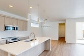 This two-story home is designed with a clean, modern aesthetic characterized by white walls, light wood-tone cabinetry, and consistent use of light hardwood flooring on the main level. The first floor hosts the open-concept kitchen and living area, where a large central island with a white quartz countertop and undermount sink serves as the focal point. Light wood flat-panel cabinetry lines the kitchen walls alongside stainless steel appliances, while pendant lighting adds a contemporary touch overhead. A sliding glass door connects the living area to a fenced outdoor space, filling the room with natural light. A compact half bathroom on the main floor features a pedestal sink and a tall vertical mirror. The second floor accommodates five bedrooms, including a primary bedroom, connected by hallways and served by two full bathrooms. The full bathrooms feature light wood vanities with quartz countertops and tiled tub-shower combinations with windows for natural ventilation. Bedrooms are carpeted and receive natural light through multiple windows, with some offering outdoor views. A dedicated closet provides additional storage. The overall ambiance is bright, neutral, and move-in ready.