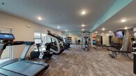 This single-floor fitness center is designed to accommodate a comprehensive range of workout routines. The space is thoughtfully laid out, with distinct zones dedicated to different types of exercise. The cardio area is well-stocked with multiple treadmills, ellipticals, stair climbers, and stationary bikes—including spin bikes—each equipped with digital screens for interactive workout experiences. Large windows line the perimeter, allowing ample natural light to fill the space and providing outdoor views that enhance the overall workout environment.

The strength training zone features a multi-station cable machine, a dedicated bench press setup, and an organized rack of medicine balls and kettlebells in various sizes, catering to both free-weight and machine-based training. The flooring transitions between durable carpet-style material and hard surface areas, clearly delineating functional zones.

A standout interior feature is the vibrant, multicolored accent wall panel above a built-in refreshment station, which includes a sink, cabinetry, and a water dispenser—adding both visual interest and practical convenience. Recessed lighting throughout, combined with a teal ceiling accent, gives the space a modern and energetic ambiance well-suited for an active fitness environment.