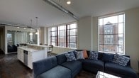 This single-floor urban loft showcases a thoughtfully designed open-concept layout that blends industrial and contemporary aesthetics. The living area is anchored by large, multi-pane windows with dark metal frames that flood the space with natural light and offer views of the surrounding cityscape. Exposed metal ductwork runs along the ceiling, adding an industrial character that complements the modern design.

The living space features dark hardwood-style flooring that provides a warm contrast to the neutral wall tones. An open kitchen is seamlessly integrated into the layout, outfitted with white cabinetry, granite countertops, and pendant lighting that hangs above a breakfast bar with seating. The kitchen flows naturally into the living area, making the overall space feel connected and functional.

The vaulted ceiling height adds a sense of openness and volume throughout. The overall ambiance strikes a balance between industrial edge and modern comfort, with clean lines, quality finishes, and an abundance of natural light creating a welcoming and stylish environment. The large windows frame urban rooftop views, reinforcing the loft's city-dwelling character throughout.