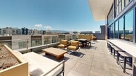Located at 553 W 100 S in Salt Lake City's Gateway district, this stylish communal lounge space at Alta Stone at the Gateway is designed to impress. The single-floor living area is anchored by soaring double-height ceilings that amplify the sense of openness and grandeur. A wall-mounted fireplace framed in textured stone tile serves as a striking focal point, complemented by a large flat-screen display above it. Plush, channel-tufted leather sectional seating and sleek black leather accent chairs with gold-toned frames create an inviting yet sophisticated atmosphere. Curated contemporary artwork adorns the walls, adding vibrant character to the neutral palette of light hardwood flooring and crisp white shiplap paneling. Floor-to-ceiling steel-framed windows and glass doors flood the interior with natural light while offering direct access to an expansive rooftop terrace. The outdoor space is thoughtfully appointed with multiple seating areas, dining tables, string lights, and raised planters — all framed by breathtaking 360-degree views of the Salt Lake City skyline and the surrounding Wasatch Mountain range. The entry features frosted glass doors and a bold branded area rug, setting a polished, welcoming tone from the moment of arrival.