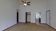 This single-story space makes a strong first impression with its dramatically vaulted ceilings, which lend an airy, open quality to the main living area. A ceiling fan with a light fixture is centrally mounted overhead, providing both airflow and ambient lighting suited to the generous vertical scale of the room. The floors are covered in neutral-toned carpet that extends throughout, complementing the clean white walls and warm wood-toned baseboards and trim that frame each area cohesively.

A built-in closet with a hanging rod is recessed into one wall, offering convenient storage directly within the main living space. To one side, an open doorway leads into a naturally lit room featuring a window with views of surrounding greenery, bringing the outdoors in and filling the space with natural light. Adjacent to this, a passage opens into what appears to be a kitchen, visible cabinetry suggesting functional prep and storage space.

A hallway on the opposite side provides access to additional areas of the home. The overall layout feels connected and navigable, with an open flow between rooms. The combination of high ceilings, neutral finishes, and natural light creates a bright, welcoming atmosphere throughout.