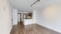 This single-floor space showcases a clean, modern design with an open-concept layout that seamlessly connects the living area and kitchen. The kitchen is well-appointed with warm taupe cabinetry, a decorative tile backsplash, stainless steel appliances including a full-size refrigerator and built-in microwave, and a crisp white kitchen island with an integrated sink. The island serves as both a functional workspace and a natural divider between the kitchen and the open living area. Warm hardwood-style flooring runs throughout the space, complementing the neutral white walls and contributing to a bright, cohesive aesthetic. Track lighting along the ceiling provides adjustable illumination, while recessed lighting adds to the overall brightness of the space. The ceilings are smooth and contemporary, enhancing the clean lines of the interior. The entryway features a dark-toned door that provides a subtle contrast to the otherwise light and airy palette. The overall ambiance is modern, minimalist, and move-in ready, offering a versatile canvas well-suited for a variety of interior styles.