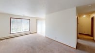 Located at 2601 S Williams Ave in Sioux Falls, South Dakota, this single-floor unit offers a functional and straightforward layout designed for comfortable everyday living. The floor plan, as revealed by the overhead scan, includes a spacious living area, a dedicated kitchen, at least one bedroom, and a full bathroom, all arranged in a practical configuration.

The living area is generously proportioned, featuring neutral wall-to-wall carpeting, white walls, and a textured ceiling. Large windows dressed with vertical blinds allow ample natural light to fill the space, while a sliding glass door provides additional brightness and potential outdoor access. Baseboard heating units run along the exterior walls, ensuring efficient warmth throughout the colder Sioux Falls seasons.

The galley-style kitchen is well-equipped with warm oak cabinetry offering abundant storage, light countertops, tile flooring, and white appliances including an electric range, microwave, dishwasher, and refrigerator. A skylight-style ceiling fixture brightens the workspace effectively.

The bedroom is a comfortable size with carpeting, a large wood-framed window overlooking surrounding greenery, and baseboard heating. The bathroom features a clean, white tub-shower combination with chrome fixtures and safety grab bars. Throughout, wood-toned trim and door accents provide warm contrast to the neutral palette.