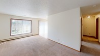 Located at 2601 S Williams Ave in Sioux Falls, South Dakota, this single-floor unit offers a functional and straightforward layout designed for comfortable everyday living. The floor plan, as revealed by the overhead scan, includes a spacious living area, a dedicated kitchen, at least one bedroom, and a full bathroom, all arranged in a practical configuration.

The living area is generously proportioned, featuring neutral wall-to-wall carpeting, white walls, and a textured ceiling. Large windows dressed with vertical blinds allow ample natural light to fill the space, while a sliding glass door provides additional brightness and potential outdoor access. Baseboard heating units run along the exterior walls, ensuring efficient warmth throughout the colder Sioux Falls seasons.

The galley-style kitchen is well-equipped with warm oak cabinetry offering abundant storage, light countertops, tile flooring, and white appliances including an electric range, microwave, dishwasher, and refrigerator. A skylight-style ceiling fixture brightens the workspace effectively.

The bedroom is a comfortable size with carpeting, a large wood-framed window overlooking surrounding greenery, and baseboard heating. The bathroom features a clean, white tub-shower combination with chrome fixtures and safety grab bars. Throughout, wood-toned trim and door accents provide warm contrast to the neutral palette.