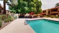 Located at 9415 N 99th Ave in Peoria, Arizona, this outdoor amenity space showcases a generously sized swimming pool as its central feature. The pool's clear, turquoise water is complemented by a wide surrounding deck that provides ample space for lounging and relaxation. Multiple seating areas are arranged around the pool, each shaded by vibrant red market umbrellas paired with dining-height tables and chairs, offering comfortable spaces for outdoor gatherings. Additional chaise lounge chairs are positioned poolside for sunbathing. The pool is accessible via stainless steel handrails and entry steps.

The space is framed by lush, mature trees that provide natural shade and a sense of seclusion, creating a pleasant contrast to the open Arizona sky above. Manicured landscaping with desert-adapted plantings borders the perimeter, enhancing the resort-like atmosphere. A gated fenced section offers added safety and separation within the outdoor area.

The backdrop features a multi-story building with a distinctive Southwestern architectural style, characterized by warm earth-tone stucco exteriors, red-tiled rooflines, and structured balconies. The overall ambiance is that of a well-maintained, inviting outdoor retreat designed for leisure and community enjoyment in the heart of the Sonoran Desert region.