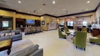 Located at 15400 Lookout Rd in Live Oak, Texas, this single-floor community clubhouse or leasing center offers a well-appointed, open-concept interior designed for both comfort and function. The space is characterized by light tile flooring, warm earth-toned walls accented with rich dark wood trim, and recessed lighting throughout, creating a welcoming and polished atmosphere.

The main gathering area features multiple seating arrangements, including plush gray sofas, accent chairs in green and striped upholstery, and a central coffee table — all arranged to encourage socializing. A dedicated lounge alcove with wraparound windows offers panoramic views of the outdoor pool and landscaped grounds, bathing the space in natural light.

A bar-height counter with seating and dark wood cabinetry serves as a media and refreshment area, complete with a mounted flat-screen TV. Adjacent spaces provide formal and casual dining options, including round tables with upholstered chairs and bar-height bistro tables.

The entrance foyer makes a strong first impression with tall ceilings, an elegant chandelier, French doors, and a built-in workstation area suitable for leasing or administrative use. A transitional room near the entrance features a patterned area rug, mixed wood tones, and direct outdoor access, rounding out this versatile and thoughtfully designed community space.