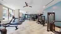 This expansive fitness center is thoughtfully designed to accommodate a wide range of workout styles and training needs. The space features a clean, modern aesthetic defined by teal accent walls, neutral patterned flooring, and recessed lighting complemented by ceiling fans throughout, ensuring comfort during intense sessions.

The cardio area lines one side of the room with multiple treadmills and step machines, positioned alongside large windows that invite natural light and provide outdoor views, creating an open and energizing atmosphere. Adjacent to this area, a dedicated cycling zone houses several screen-equipped stationary bikes, ideal for interactive or instructor-led workouts.

A standout feature is the turf sprint lane marked with yardage lines, offering space for functional training drills and athletic conditioning. Surrounding this central lane is a comprehensive strength training section, including power racks, cable machines, a full dumbbell rack with adjustable benches, and a TRX suspension system. Full-length mirrors along the walls enhance the sense of space while supporting proper form.

Near the entrance, a small refreshment station with a mini-fridge and counter space adds a practical amenity. Glass-paned doors connect the fitness center to the broader facility, reinforcing its accessible and community-oriented design.