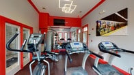 Located at 750 Boone Station Dr in Burlington, NC, this single-floor fitness center is designed to deliver a fully equipped workout experience in a visually energetic environment. The space is defined by a striking red-and-gray color scheme, with bold red accent walls, trim, and door frames contrasting against light gray walls and a white ceiling — creating a high-energy atmosphere conducive to exercise.

The facility is outfitted with a comprehensive selection of True-brand cardio machines, including multiple treadmills and elliptical cross-trainers, positioned to take advantage of the natural light streaming through several red-framed windows. A full dumbbell rack with a range of free weights, a cable strength machine with an adjustable bench, and additional resistance equipment round out the strength training options.

Large wall-mounted mirrors expand the perceived space and allow users to monitor their form. Overhead, modern geometric LED light fixtures provide bright, even illumination throughout. A wall-mounted flat-screen television adds an entertainment element to cardio sessions. Rubberized flooring covers the workout area, providing both safety and durability. Vibrant wall art adds a lively, urban aesthetic touch. Glass-paned doors offer views of and access to an adjacent outdoor area, allowing natural light to further enhance the space.