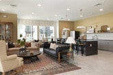 This clubhouse in Idaho Falls, Idaho, offers a thoughtfully designed single-floor layout that seamlessly blends comfort and functionality. The open-concept floor plan connects the living and kitchen areas, creating a spacious, inviting atmosphere ideal for gathering and socializing.

The living room is a standout centerpiece, featuring a sleek wall-mounted fireplace framed by white shiplap paneling and a large flat-screen TV above. Multiple seating arrangements — including plush sofas, leather tufted couches, and accent armchairs — are anchored by a rich, patterned area rug atop gleaming large-format tile flooring. Abundant natural light pours in through numerous windows dressed with neutral curtains and blinds, while recessed lighting provides warm, even illumination throughout. Bold geometric wallpaper accents add a modern touch to select walls.

The kitchen is stylishly appointed with gray cabinetry, dark granite countertops, white subway tile backsplash, and warm brass hardware and fixtures. A French door refrigerator in white with bronze accents serves as a striking focal point. Adjacent to the kitchen, a built-in banquette dining nook with a round table and pendant lighting offers a charming spot for casual meals or conversation. Together, these spaces create a warm, well-equipped community hub.