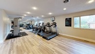 This single-level fitness center offers a comprehensive workout environment designed to accommodate a variety of exercise routines. The space is organized into distinct functional zones, each dedicated to a specific type of training. Light hardwood-style flooring runs throughout, lending a clean, modern aesthetic that contrasts with the protective rubber mats placed beneath the equipment.

The cardio section features a row of treadmills and elliptical machines aligned along one wall, positioned near windows that allow natural light to filter into the space. A large mirror spans a portion of the wall, visually expanding the room and allowing users to monitor their form during exercise.

The strength training area is anchored by a Cybex functional trainer, a versatile cable machine mounted on a protective rubber mat. Adjacent to this station, stability balls in multiple sizes are displayed on a dedicated rack, alongside wall-mounted exercise mats and a flat-screen television.

Recessed lighting and textured ceilings contribute to a bright, well-lit atmosphere throughout. Posted fitness center rules indicate this is a shared or community facility. The overall layout is spacious and thoughtfully arranged, offering an accessible and functional environment for diverse fitness needs.