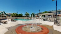 Located in West Jordan, Utah, this community recreational facility offers an impressive array of outdoor amenities designed for relaxation and leisure. At the heart of the space is a generously sized outdoor swimming pool with crystal-clear turquoise water, accessible via stainless steel entry rails and surrounded by a wide concrete pool deck. The deck is furnished with numerous lounge chairs, providing ample space for sunbathing under Utah's abundant sunshine.

A dedicated splash pad area sits adjacent to the pool zone, enclosed by white vinyl fencing and illuminated by decorative post lights — an ideal feature for younger visitors. The pergola structure on one side of the deck offers shaded seating and an inviting gathering spot.

The on-site clubhouse is a standout architectural element, featuring a distinctive green pitched roof, stone and stucco exterior, and expansive floor-to-ceiling windows that allow natural light to fill the interior while offering views of the pool area. The surrounding backdrop includes mature trees and the scenic mountain range visible on the horizon, enhancing the overall ambiance of the space.

The entire recreational area is enclosed by white vinyl privacy fencing with gated access points. Tall pole lighting throughout ensures the space is well-lit for evening use. This well-appointed outdoor community space blends functional amenities with an inviting, resort-like atmosphere.