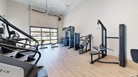 This fitness center at 4170-4190 E Commerce Way in Sacramento, California, offers a well-appointed exercise space designed for a comprehensive workout experience. The room features soaring ceilings that lend an open, airy feel, enhanced by recessed lighting and a large ceiling fan that promotes airflow throughout the space. Light wood-look flooring runs throughout, providing a clean and modern aesthetic.

The exercise room is thoughtfully organized to accommodate a wide range of fitness activities. Cardio equipment includes treadmills, an elliptical trainer, and a stair climber, all positioned to take advantage of the natural light. A wall-mounted flat-screen television adds convenience for those exercising. The strength training area is equally well-stocked, featuring cable machines, a multi-functional weight station, a dumbbell rack, and adjustable benches.

A standout architectural feature is the full-width glass garage-style door that opens to outdoor green space, flooding the interior with natural light and providing refreshing views of the surrounding landscape. Floor-to-ceiling glass partition walls with sleek black framing further enhance the bright, transparent feel of the space. A wall-mounted speaker system adds to the amenity. This fitness center delivers a bright, modern, and fully functional environment suited for a variety of workout routines.