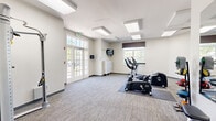 This fitness center in Windsor Mill, MD, is a well-appointed, single-floor facility designed to support a range of workout needs. The space features neutral-toned walls, textured carpet tile flooring, and flush-panel ceiling light fixtures that provide consistent, even illumination throughout. Multiple windows, including a set of French doors, allow natural light to fill the main exercise area, creating an open and welcoming atmosphere with views of the surrounding outdoor environment.

The main workout area is equipped with a variety of cardio machines, including treadmills, ellipticals, and stationary bikes from brands such as Cybex. A cable-based strength machine offers versatile resistance training options, while a weight bench and medicine ball rack provide additional tools for functional fitness. A wall-mounted television is positioned for viewing during workouts.

A dedicated secondary area features a large wall mirror, a weight bench, and a foam roller station, offering space for stretching, strength exercises, and warm-up routines. A paper towel dispenser and sanitation station are conveniently accessible. Restroom facilities are accessible directly from this area. Security is supported by a mounted camera. Overall, the fitness center presents a clean, functional, and organized environment suited for a variety of exercise routines.