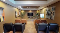 This single-floor lounge space is thoughtfully designed with a warm, contemporary aesthetic that blends comfort with sophistication. The main lounge area centers around a wall-mounted flat-screen television flanked by tall, lush indoor palms, creating a focal point that draws the eye immediately upon entry. Seating is arranged in a structured layout, featuring a mix of tan and black leather armchairs that provide ample accommodations for guests or residents.

The flooring throughout is a wood-look plank surface, adding a warm, cohesive feel to the space. The ceilings feature a distinctive tiered tray design with built-in and track lighting, lending architectural depth and allowing for flexible, layered illumination. Warm earth tones — ranging from golden ochre to deep taupe — coat the walls and complement the overall interior palette.

Curated wall art, including pop-culture inspired prints and abstract canvases, adds personality and visual interest throughout the room. On one end, windows fitted with honeycomb shades allow natural light to filter in while offering views of an outdoor pool area. An adjacent room, visible through a wide doorway, reveals a bar or lounge counter with bar-height seating, suggesting a multifunctional entertainment environment. The overall ambiance is inviting, polished, and well-suited for social gatherings or relaxed lounging.