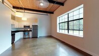 This 1-bedroom, 1-bathroom unit showcases a compelling industrial-chic aesthetic rooted in raw, architectural materials and a thoughtfully open layout. Polished concrete floors run throughout the entire space, reflecting natural light and lending a clean, urban character. Overhead, warm wood-plank ceilings are intersected by bold, exposed steel beams, creating a striking visual contrast that defines the loft-style ambiance.

The open living area is anchored by a large, multi-pane window with a black steel frame, offering generous views of the surrounding greenery and flooding the room with daylight. Pendant drum lights suspended from the ceiling provide warm, focused illumination throughout.

The kitchen is efficiently designed with white cabinetry, dark granite countertops, a light blue tile backsplash, and a full suite of stainless steel appliances, including a refrigerator, electric range, microwave, and dishwasher. Ample upper and lower cabinet storage ensures strong functionality.

The bathroom features a clean, minimalist design with white walls, a full tub-shower combination, and polished concrete flooring consistent with the rest of the unit. The bedroom offers a serene retreat, highlighted by the same large grid-pane window and soaring wood-and-beam ceiling that define the overall character of this distinctive space.