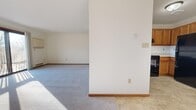 This single-floor residence offers a clean, functional layout designed to maximize comfort and livability. The spacious main living area is carpeted in neutral gray and bathed in natural light streaming through a sliding glass door that opens directly onto a wood-railed outdoor deck, providing a seamless connection to the exterior. Baseboard heating runs along the perimeter of the living space, ensuring efficient warmth throughout. A partial wall divides the living area from the kitchen, maintaining an open feel while defining each zone. The kitchen features warm oak cabinetry with ample upper and lower storage, light-toned tile flooring, and black appliances including a refrigerator and range. A dedicated bedroom offers plush carpeting, a large window with exterior views, and baseboard heating. The bathroom includes a full tub-and-shower combination with clean white surroundings and tile flooring. A wood-banistered staircase and landing area add a classic residential touch to the interior. Throughout the space, white walls and consistent trim work in warm wood tones create a cohesive, understated aesthetic well-suited for a variety of living arrangements.
