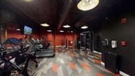 This single-floor fitness center at Canal Bank Road in Windsor Locks, Connecticut, delivers a well-equipped workout environment defined by a striking industrial aesthetic. The space is characterized by a dark, exposed ceiling with visible ductwork and structural beams, creating a loft-like atmosphere with generous vertical clearance. Original wood columns punctuate the floor plan, adding a raw, architectural character that complements the bold design palette.

The walls are painted in deep red and matte black, establishing an energetic and focused atmosphere throughout. Underfoot, a distinctive geometric tile pattern in charcoal, gray, and orange adds visual dynamism and reinforces the space's high-energy design theme. Overhead, large dome-shaped pendant lights provide warm, even illumination across the entire area.

The fitness floor is comprehensively equipped with a row of treadmills and elliptical trainers along one side, complemented by spin bikes positioned in front of full-length mirrors and wall-mounted televisions for interactive workout experiences. The strength training zone includes cable machines, a functional trainer, a seated press station, and a dumbbell rack. A water fountain and sanitizing station are conveniently located near the entrance, and motivational text is displayed near the doorway, reinforcing the space's purpose-driven design.