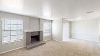 Located at 8305 Centaur Ct in Fort Worth, TX, this two-story home offers a thoughtfully designed layout with a clean, neutral aesthetic throughout. The main living area is a generously sized, carpeted space anchored by a classic brick fireplace with a raised hearth and a white mantel, flanked by multiple windows fitted with blinds that allow natural light to fill the room. The textured ceiling and soft wall tones contribute to a warm, inviting atmosphere.

The kitchen is well-appointed with gray cabinetry, white countertops, and a crisp white subway tile backsplash. Stainless steel appliances—including a range, microwave, and refrigerator—add a modern touch, while pendant lighting above the sink area enhances the functional charm. Dark hardwood-style flooring grounds the kitchen space.

A staircase leads to the upper level, where carpeted bedrooms continue the home's consistent neutral palette. At least one bedroom features vaulted ceilings with a ceiling fan and a bright window. The bathroom includes a tub and shower combination with large-format stone-look tile surround and chrome fixtures, providing a clean and updated finish. Additional carpeted rooms offer flexible living or sleeping arrangements throughout the home.