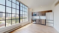 This thoughtfully designed single-floor unit offers a contemporary living experience defined by clean lines, modern finishes, and abundant natural light. The most striking architectural feature is the expansive floor-to-ceiling grid-style windows in the main living area, which flood the space with sunlight and frame elevated views of the surrounding cityscape and treetops, lending the unit an airy, loft-like quality.

The open kitchen is efficiently appointed with warm wood-toned cabinetry accented by matte black hardware, white subway tile backsplash, light stone countertops, and a full suite of stainless steel appliances including a gas range, over-the-range microwave, dishwasher, and a multi-door refrigerator. Light hardwood-style flooring runs consistently throughout the living areas and bedroom, contributing to a cohesive, modern aesthetic.

A hallway connects the main living space to the private bedroom, which features double closet doors with a clean white panel design and black hardware. The full bathroom is finished with large-format neutral tile flooring, a frameless sliding glass shower enclosure with white tile surround, and a wood-accented vanity. Recessed lighting and crisp white walls throughout the unit reinforce its modern, minimalist character.