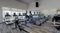 This single-floor fitness center offers a comprehensive and thoughtfully designed workout environment. The space is anchored by an impressive selection of cardio equipment, including treadmills, elliptical machines, stair climbers, and a rowing machine — all arranged in organized rows to maximize usability and flow. Full-length mirrors line one wall, enhancing the sense of openness while allowing members to monitor their form during exercise. Large floor-to-ceiling windows flood the space with abundant natural light and provide views of the surrounding outdoor area, creating an inviting and energizing atmosphere. The elevated ceiling height adds to the spacious feel of the room, preventing any sense of crowding even with the generous equipment offerings. Striped, durable flooring runs throughout, adding visual depth while standing up to the demands of daily use. Wall-mounted speakers ensure an immersive audio experience during workouts. The clean, neutral color palette and bright, modern finishes contribute to a polished, professional ambiance, making this fitness center a well-rounded facility suited for a variety of cardio training needs.