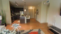This well-appointed single-floor residence showcases a thoughtfully designed open-concept layout that seamlessly connects the main living areas. Warm hardwood flooring flows throughout the space, creating a cohesive and inviting atmosphere from room to room.

The kitchen is a standout feature, outfitted with rich dark cabinetry, stainless steel appliances, and a granite-topped island that doubles as a casual dining and prep area. Pendant lighting in a warm red hue hangs above the island, adding a contemporary decorative touch. Track lighting overhead ensures the kitchen remains bright and functional.

The kitchen island transitions naturally into a dining area, where white upholstered chairs complement the dark tones of the cabinetry and island base. An adjacent entryway features a paneled door and a small closet, providing practical storage and a welcoming entry point.

The living area, partially visible in the foreground, is anchored by a bold patterned area rug in grey and red, adding a vibrant focal point to the space. Neutral, light-toned walls throughout the residence create a clean backdrop that allows furnishings and finishes to stand out. The overall ambiance is modern, warm, and functional — ideal for both everyday living and casual entertaining.