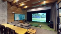 This single-level space is designed as a dedicated indoor golf simulator room, combining entertainment and leisure in a thoughtfully curated environment. The room's most striking feature is the large high-definition projection screen dominating one wall, creating an immersive golf simulation experience complete with a professional-grade hitting mat and artificial turf zone positioned directly in front of it. Warm wood paneling lines the walls and carries through to the exposed beam ceiling, giving the space a rich, cabin-like aesthetic that balances functionality with comfort. Recessed ceiling lighting provides even, well-controlled illumination throughout. A secondary flat-screen display is mounted on the adjacent wall, offering additional viewing options. The lounge area is furnished with generous sectional seating and bean bag chairs, inviting spectators to relax while others play. A rustic wooden dining or activity table with matching bench seating anchors the social area of the room. The combination of natural wood tones, green turf, and modern technology creates a cohesive ambiance that is both sporty and sophisticated, making this space ideal for golf enthusiasts and entertainment alike.