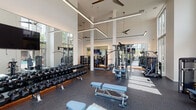 This well-appointed fitness center, branded "SWEAT," delivers a premium workout experience through thoughtful design and a comprehensive selection of equipment. The space is defined by soaring double-height ceilings, floor-to-ceiling glass windows, and abundant natural light that floods the interior throughout the day, offering views of lush palm trees and the surrounding outdoor amenity areas, including a pool.

Upon entry, a sculptural white accent wall featuring geometric cutouts sets a modern tone, leading into the expansive main workout floor. Linear LED lighting elements are suspended from the high ceilings, complementing the sleek, all-white architectural palette. Ceiling fans provide additional airflow across the open floor plan.

The main exercise area is outfitted with a full rack of dumbbells, adjustable benches, and multiple cable and strength machines. A dedicated cardio zone features a row of treadmills and elliptical trainers positioned directly in front of floor-to-ceiling windows. A Peloton station with stationary bikes is also available. Large wall-mounted mirrors expand the sense of space and allow users to monitor their form throughout their workout. Durable rubber flooring covers the entire space, ensuring safety and comfort. The overall ambiance is clean, energizing, and professionally designed.