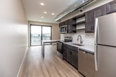 This modern single-floor unit offers a well-designed open-concept layout that seamlessly connects the kitchen and living area. The kitchen is a standout feature, showcasing dark wood cabinetry, light quartz countertops, and a classic white subway tile backsplash. Stainless steel appliances — including a refrigerator, electric range, over-the-range microwave, and dishwasher — are complemented by open shelving and a stainless undermount sink. An exposed metallic HVAC duct runs through the ceiling, lending an industrial character to the space. Light wood-tone flooring extends throughout the unit, creating a warm, cohesive aesthetic. The living area benefits from abundant natural light and direct access to a private balcony through floor-to-ceiling sliding glass doors with black frames, offering expansive views of the surrounding cityscape, including a Ferris wheel and bridge in the distance. The bathroom features a dark wood vanity with a white countertop, complemented by clean white walls and matching flooring. A convenient in-unit laundry area accommodates a stacked washer and dryer. Recessed lighting throughout ensures a bright and inviting atmosphere across all areas of the space.