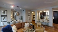 This single-floor space showcases an open-concept layout that seamlessly connects the living area to the kitchen, creating a cohesive and functional environment. The living area is anchored by a rich brown leather sofa accented with decorative pillows, complemented by a modern armchair and a dark coffee table. A distinctive oversized metallic globe pendant lamp and track lighting overhead provide both ambient and accent illumination, contributing to the warm, inviting atmosphere.

Hardwood flooring runs throughout, adding warmth and continuity across the space. Neutral gray walls serve as a versatile backdrop for the carefully curated décor, including framed artwork and lush indoor greenery.

The kitchen, visible from the living area, features dark cabinetry, stainless steel appliances including a built-in microwave and oven, and a breakfast bar with modern white counter-height chairs. A sleek pendant light hangs above the bar, adding a refined touch. A hallway connecting the spaces hints at additional rooms beyond, while a bold geometric art piece near the entry adds a striking visual element. The overall ambiance is modern, warm, and well-appointed.