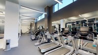 This contemporary fitness center offers a thoughtfully designed workout environment that balances functionality with a sleek, modern aesthetic. The space features impressively high ceilings with clerestory windows that flood the interior with abundant natural light, creating an open and energizing atmosphere. Linear LED lighting runs across the ceiling, ensuring consistent, well-distributed illumination throughout the facility. The main workout floor is generously proportioned, providing ample space between equipment for comfortable, unobstructed movement. A full row of cardio machines — including treadmills, ellipticals, and stair climbers — lines the floor, all equipped with modern digital displays. Structural columns with a dark finish add a bold architectural contrast to the lighter walls and ceiling. Full-length mirrors along the walls expand the sense of space while allowing users to monitor their form. A dedicated strength training and free weights area is visible in the background, rounding out the facility's comprehensive offerings. The dark, rubberized flooring provides durability and safety underfoot. Overall, the space exudes a professional, polished character that caters to a wide range of fitness needs in a bright and inviting setting.