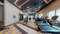 This fitness center combines contemporary design with comprehensive amenities to create a fully functional workout facility. The space is defined by its striking dark ceiling adorned with geometric LED light strips arranged in angular, hexagonal patterns, complemented by recessed lighting and ceiling fans that maintain a comfortable atmosphere throughout.

The main workout floor is spaciously laid out and features a distinctive patterned carpet in gold, grey, and charcoal tones that delineates different workout zones. The cardio area is equipped with a row of Star Trac treadmills and elliptical machines positioned along large windows that invite natural light and outdoor views. A central strength training zone houses a robust multi-functional cable and functional training rig by Hoist, surrounded by a full dumbbell rack and a variety of resistance machines.

Large mirrors along the walls enhance the sense of space and support training visibility. A dedicated entrance area includes built-in shelving, a service counter, hand sanitizing station, and glass-paneled doors, providing an organized and welcoming transition into the facility. The overall design balances aesthetic sophistication with practical functionality.