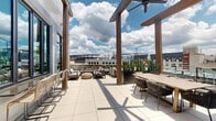 Located at 2801 Windy Ridge Pkwy SE in Smyrna, Georgia, this rooftop amenity deck offers a sophisticated blend of covered and open-air spaces designed for socializing and relaxation. The expansive terrace is finished with large-format porcelain tile throughout, lending a clean, modern aesthetic that contrasts beautifully with the warm wood structural elements.

The covered portion of the deck features an exposed wood-beam ceiling supported by substantial timber columns, ceiling fans for comfort, and wall-mounted televisions. This sheltered area accommodates a full-size shuffleboard table, multiple dining tables with rope-woven chairs, and a built-in cabinetry unit. Exposed brick accent walls add texture and warmth to this semi-enclosed gathering space.

The open-air section of the terrace extends outward beneath a wood pergola structure, offering unobstructed panoramic views of the Smyrna and greater Atlanta skyline. Lounge seating with plush cushions, planters featuring tall snake plants, and bistro-style tables create an inviting outdoor living environment. Glass railings along the perimeter ensure safety while preserving the expansive sightlines.

Together, the covered and open zones create a versatile, well-appointed rooftop destination that balances urban energy with relaxed outdoor comfort.