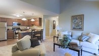 This single-floor residence offers an inviting open-concept design that effortlessly blends the living, dining, and kitchen areas into one cohesive and functional space. The living room is anchored by plush, light-toned seating arranged around a dark wood coffee table, creating a relaxed and comfortable atmosphere. Soft carpeting extends across the floor, adding warmth underfoot, while the soaring vaulted ceiling amplifies the sense of openness and airiness throughout the main living area. A framed piece of wall art adds a tasteful decorative accent to the space. Adjacent to the living room, the dining area features a dark bistro-style table and chairs, positioned beneath elegant pendant lighting that casts a warm, focused glow. The kitchen is finished with rich warm-toned cabinetry and a dark countertop, providing ample workspace and storage. Pendant lights above the kitchen bar area echo the dining fixtures, tying the spaces together visually. The overall color palette—soft greens, beiges, and warm browns—creates a calm, neutral ambiance throughout. A hallway visible from the main space suggests additional private rooms beyond, completing this well-appointed and thoughtfully designed home.