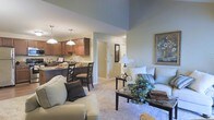 This single-floor residence offers an inviting open-concept design that effortlessly blends the living, dining, and kitchen areas into one cohesive and functional space. The living room is anchored by plush, light-toned seating arranged around a dark wood coffee table, creating a relaxed and comfortable atmosphere. Soft carpeting extends across the floor, adding warmth underfoot, while the soaring vaulted ceiling amplifies the sense of openness and airiness throughout the main living area. A framed piece of wall art adds a tasteful decorative accent to the space. Adjacent to the living room, the dining area features a dark bistro-style table and chairs, positioned beneath elegant pendant lighting that casts a warm, focused glow. The kitchen is finished with rich warm-toned cabinetry and a dark countertop, providing ample workspace and storage. Pendant lights above the kitchen bar area echo the dining fixtures, tying the spaces together visually. The overall color palette—soft greens, beiges, and warm browns—creates a calm, neutral ambiance throughout. A hallway visible from the main space suggests additional private rooms beyond, completing this well-appointed and thoughtfully designed home.