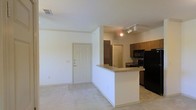 Located in Roanoke, TX, this single-story residence offers a practical and comfortable open-concept floor plan. The living area is finished with light carpeting, creating a neutral and welcoming atmosphere that flows seamlessly into the kitchen and dining space.

The kitchen is a central feature of the home, equipped with black appliances including a full-size refrigerator and range, and accented by a tasteful tile backsplash. Warm-toned wood cabinetry provides generous storage, while the peninsula-style breakfast bar offers additional counter space and serves as a natural dividing point between the kitchen and living area. Track lighting above the kitchen ensures bright, functional illumination throughout the cooking space.

The entryway is marked by a classic paneled door, adding a traditional touch to the home's clean, simple aesthetic. The light-colored walls throughout enhance the sense of openness and reflect natural light effectively. The overall layout is well-suited for everyday living, offering a connected and efficient use of space across the main living and kitchen areas. The single-floor design makes the residence easily navigable, with a straightforward flow between spaces.