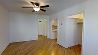 This single-floor residence showcases a clean, functional layout with warm hardwood flooring that runs throughout the main living area, lending a sense of continuity and natural warmth to the space. The living room is anchored by a ceiling fan with an integrated light fixture, providing both airflow and ambient lighting. Crisp white walls keep the interior feeling bright and open.

A distinctive arched doorway serves as a architectural focal point, gracefully connecting the living area to the kitchen beyond. The kitchen is outfitted with white upper cabinetry, a countertop workspace, and an overhead range hood, offering a practical and tidy cooking environment. The contrast between the dark countertops and white cabinetry adds a subtle visual interest to the kitchen's design.

Adjacent to the arched passageway, a traditional hinged door with a white frame provides access to an additional area, likely a closet or secondary space, contributing to the overall storage and utility of the unit. The combination of classic architectural details, consistent flooring, and a neutral color palette creates a cohesive and inviting ambiance throughout the space.