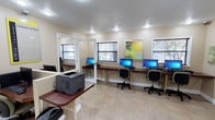 This computer lab is a single-floor space designed to provide a functional and comfortable environment for technology use. The main area features a row of desktop computer workstations lined along a rich, dark wood counter that runs along the wall, each station equipped with dual monitors and an ergonomic rolling chair. Windows fitted with horizontal blinds line the walls, allowing natural light to filter in while maintaining privacy. A vibrant piece of wall art adds a touch of color to the otherwise neutral, cream-toned interior. The flooring throughout consists of light-toned tile, contributing to the bright and clean aesthetic of the space. A dedicated printer station is conveniently positioned near the entrance, housing a laser printer atop a built-in counter for easy access. A separate enclosed alcove, accessible through a glass-paneled door, offers additional workstation space for more focused or private use. Recessed lighting provides consistent, even illumination throughout. Toward one end of the room, a casual seating area with cushioned chairs offers a relaxed spot for waiting or informal use, rounding out the lab's practical and welcoming design.