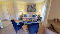 Located at 11302 10th Ave Ct E, Unit C202, in Tacoma, Washington, this single-floor residence offers a thoughtfully designed layout with a warm and inviting atmosphere. The living area serves as the heart of the home, featuring soft neutral wall tones that create a bright and airy feel. Plush carpeted flooring adds comfort underfoot, while large windows dressed in elegant curtains allow natural light to filter in, enhancing the cheerful ambiance of the room. The space is well-proportioned, comfortably accommodating a full seating arrangement that includes a generous sofa and bold navy blue accent chairs centered around a wooden coffee table. A patterned area rug anchors the seating area, adding visual depth and texture. A large abstract wall canvas serves as a striking focal point above the sofa, complemented by matching table lamps that provide warm, layered lighting throughout the space. Built-in ventilation and thoughtful architectural details contribute to the home's functional design. The overall aesthetic strikes a balance between comfort and style, making this Tacoma residence a welcoming and livable space suitable for a variety of lifestyles.