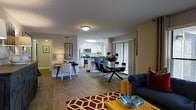This single-floor residence offers a cohesive open-concept design that seamlessly connects the main living areas. Hardwood-style flooring flows continuously throughout, lending a sense of warmth and visual continuity to the space.

The living area is anchored by a plush navy sofa and accented with a bold geometric-patterned area rug in warm red tones, creating an inviting focal point. Adjacent to the living space, a glass-topped dining table surrounded by sleek black chairs provides an elegant yet modern setting for meals and gatherings. A compact desk nook with a lamp and white chair nestles along one wall, offering a functional workspace without disrupting the open flow.

The kitchen is finished with white cabinetry, stainless steel appliances — including a range, microwave, and refrigerator — and a breakfast bar with counter seating, making it equally suited for casual dining and meal preparation. A sliding glass door off the dining area welcomes natural light and suggests access to an outdoor space beyond.

Neutral gray walls throughout keep the atmosphere calm and versatile, while thoughtful décor accents, including framed wall art and fresh floral arrangements, add personality and life to the interior. The layout is efficient, functional, and well-suited to everyday living.