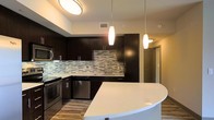 This single-floor kitchen showcases a contemporary design defined by a strong contrast between dark espresso cabinetry and bright white surfaces. The full-height dark cabinets provide generous storage, while sleek chrome hardware adds a refined, modern touch throughout. A striking mosaic tile backsplash in muted grays and whites runs the length of the cooking and prep area, serving as a visual focal point that bridges the dark and light elements of the space. Stainless steel appliances, including a range, microwave, and dishwasher, complement the kitchen's modern aesthetic. A curved peninsula with a smooth white countertop extends into the living area, offering additional prep space and a natural gathering spot. Three pendant lights — two elongated and one teardrop-shaped — hang at varying heights above the kitchen, casting warm, focused illumination that enhances the overall ambiance. A flush-mount ceiling fixture provides broader ambient lighting. The hardwood-style flooring grounds the space with a warm, natural tone that balances the boldness of the cabinetry, resulting in a cohesive and well-appointed kitchen interior.