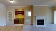 Located at 1310 Silver Sage Dr in Raleigh, NC, this single-story home offers a comfortable and functional layout designed with everyday living in mind. The main living area is anchored by a classic white-surround fireplace, providing a warm focal point for the room. Plush wall-to-wall carpeting runs throughout, adding a soft, inviting feel underfoot. Crown molding accents the ceilings, lending a touch of refined detail to the space. A standout feature of the living area is a built-in desk nook, complete with rich wood cabinetry, open shelving, and a granite-style countertop — an ideal dedicated workspace or study area seamlessly integrated into the home's design. The neutral, warm-toned walls create a versatile backdrop that complements a variety of décor styles. A doorway leads to an adjoining area, offering a glimpse of natural light filtering in through an exterior door, suggesting a well-connected and naturally lit floor plan. The overall ambiance of the home is warm, practical, and move-in ready, with thoughtful built-in details that enhance both form and function throughout the single-level layout.